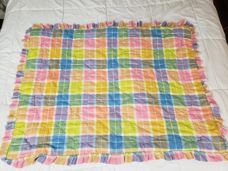 May include: A pastel plaid quilted throw blanket with a ruffled edge. The blanket is made of soft cotton and features a variety of colors, including pink, yellow, blue, and green.