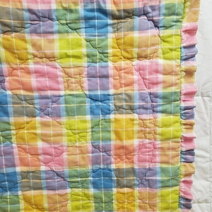 May include: A multi-colored plaid quilt with a ruffled edge. The quilt is made of a soft, cotton fabric and features a variety of pastel colors, including pink, blue, yellow, and green.