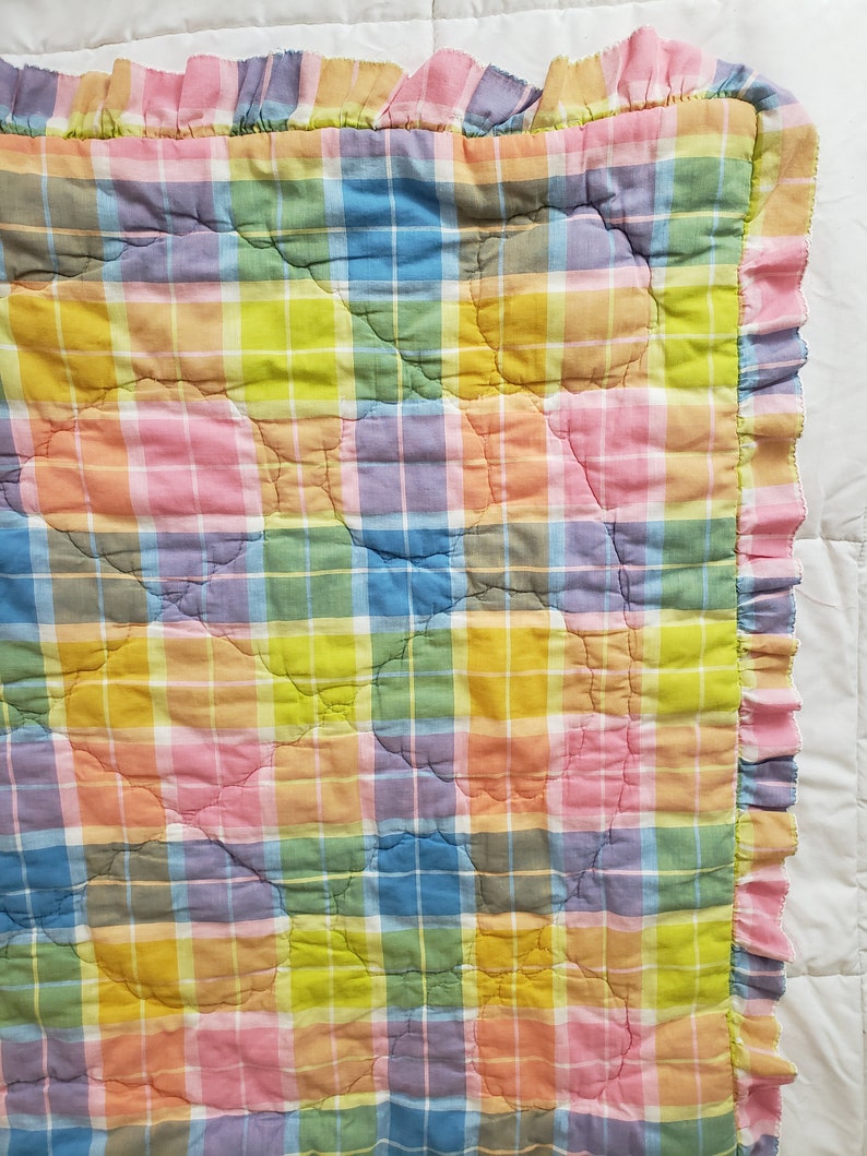 May include: A multi-colored plaid quilt with a ruffled edge. The quilt is made of a soft, cotton fabric and features a variety of pastel colors, including pink, blue, yellow, and green.