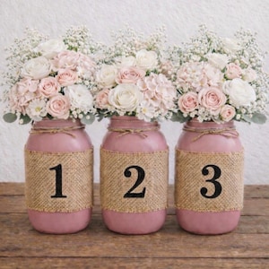 May include: Three pink mason jars with burlap accents and numbers 1, 2, and 3. Each jar holds a floral arrangement of white and pink roses, hydrangeas, and baby's breath, tied with twine. The jars sit on a wooden surface.