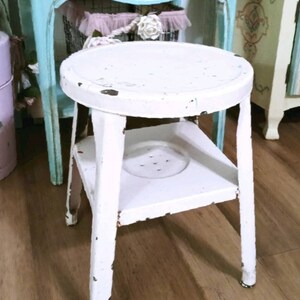 May include: White metal stool with a round top and a shelf underneath. The stool has a distressed, vintage look.
