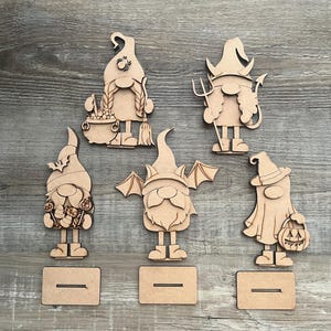 May include: Five wooden gnome cutouts with Halloween themes. Each gnome has a unique design, including a witch, devil, and vampire. Each gnome has a slot for a base. The cutouts are made of light brown wood.