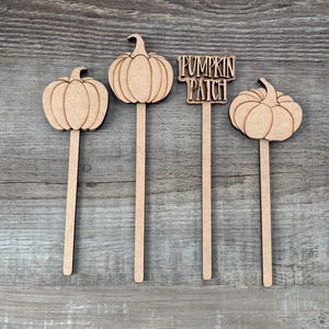 May include: Four light brown wooden plant markers featuring three pumpkin shapes and one rectangular sign with the text PUMPKIN PATCH. Each cutout is attached to a thin wooden stake, displaying a natural wood grain texture and smooth edges.