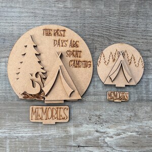 May include: Laser-cut wooden camping-themed decorations. One large piece features a tent, tree, and the text "The Best Days Are Spent Camping." A smaller piece shows a tent and trees. Includes a "Memories" sign.