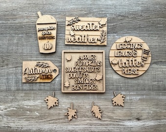 May include: A collection of laser-cut wooden fall-themed decorations. Includes a coffee cup with "pumpkin spice everything", a sign with "sweater weather", a round sign with "leggings, leaves & lattes please", and other fall-related words and leaf cutouts.