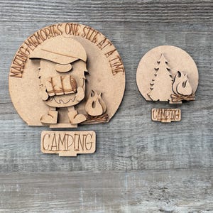May include: Wooden camping-themed decorations. One piece features a cartoon camper with the text "MAKING MEMORIES ONE SITE AT A TIME" and "CAMPING". The other piece shows a campfire and tree with the word "CAMPING".