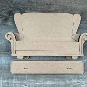May include: A wooden miniature sofa with a matching footrest, both in a light tan color. The sofa has a high back and scrolled armrests. The footrest is rectangular with two small cutouts. Perfect for dollhouses or craft projects.