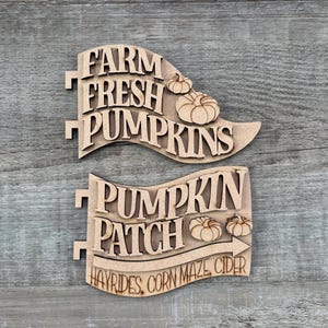 May include: Two light-colored wooden signs with laser-cut lettering. The top sign reads FARM FRESH PUMPKINS with two pumpkin illustrations. The bottom sign reads PUMPKIN PATCH with two pumpkins and the text HAYRIDES, CORN MAZE, CIDER below an arrow.