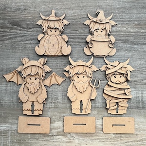 May include: Wooden Halloween-themed Highland cow cutouts. Each cutout is a different character, including a pumpkin, witch, bat, devil, and mummy. Each figure has a base for standing. The cutouts are made of light brown wood.