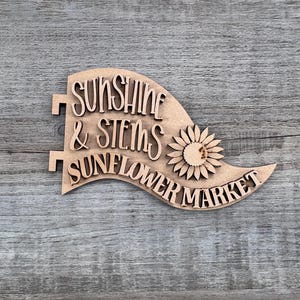 May include: A light brown wooden sign shaped like a pennant flag with the text Sunshine & Stems Sunflower Market in raised lettering. The sign features a cutout sunflower icon on the right side and two small rectangular tabs on the left edge.