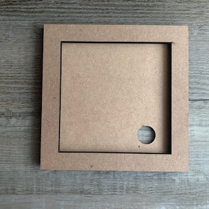 May include: A square wooden frame with a cutout in the center. The frame is brown and has a black border. There is a small round hole in the center of the cutout.