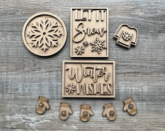 May include: Wooden craft cutouts with winter themes. The cutouts include a snowflake, a mitten, a sweater, and two signs that say "Let it Snow" and "Winter Wishes".