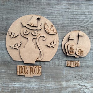 May include: Wooden Halloween decorations featuring a cat wearing a witch hat, bats, and the words "Hocus Pocus." Another decoration shows potion bottles with the words "Rat Wings" and "Bat Tails," and the word "Potions."
