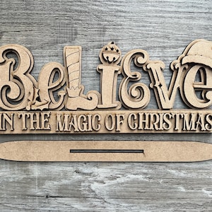 May include: Wooden sign with the words "Believe in the Magic of Christmas" in a decorative font. The sign is made of multiple pieces of wood that are layered on top of each other. The sign is painted in a light brown color.