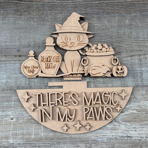May include: A laser-cut wooden sign featuring a cat wearing a witch hat, a cauldron, and potion bottles labeled Nine Lives Elixir and Black Cat Brew. Below, a semi-circular piece displays the text There's Magic In My Paws with star accents.