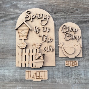 May include: Wooden spring-themed decorations. The largest piece reads "Spring is in the air" with a birdhouse, birds, and a fence. Additional pieces say "Chirp Chirp", "Tweet", and "Spring".