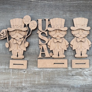 May include: Wooden Uncle Sam figures with a flag that says "USA" and balloons. The figures are wearing top hats and bow ties. The word "USA" is also spelled out in wooden letters. The figures are on wooden stands.