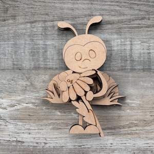 May include: A wooden laser-cut insect-like figure with a smiling face, antennae, and a flower. The figure is light brown and has a detailed design, including a daisy and grass.