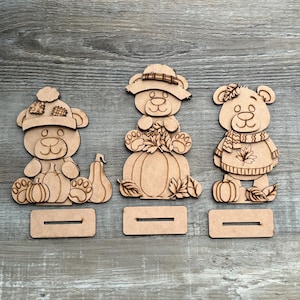 May include: Three wooden bear figurines with fall-themed designs. One bear wears a hat, another sits on a pumpkin, and the third wears a sweater and scarf. Each bear has a slot for display.