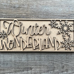 May include: A rectangular wooden sign with the words "Winter Wonderland" cut out in a cursive font. Decorative snowflakes are cut out around the edges. The sign is made of light brown wood and sits on a gray wooden surface.