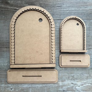 May include: Two unfinished wooden display stands. The larger stand has an arched top with a scalloped edge and a small hole. The smaller stand has a similar design. Both have a rectangular base with a slot.