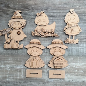 May include: A collection of wooden duck figurines in various autumn-themed outfits. One duck is a scarecrow with a sign that reads "Scarecrow On Duty." Other ducks are adorned with hats, scarves, and fall foliage. Each figurine has a slot for a stand.