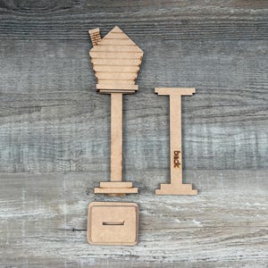May include: A wooden birdhouse kit with a house-shaped top, a post, and a base. The kit includes a separate piece with the word "back" etched on it. The pieces are made of light brown wood.