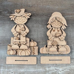 May include: Two unfinished laser-cut wooden scarecrow figures, each featuring a hat, layered clothing details, and pumpkin accents. Both figures are positioned above separate rectangular wooden bases with horizontal slots for display purposes.