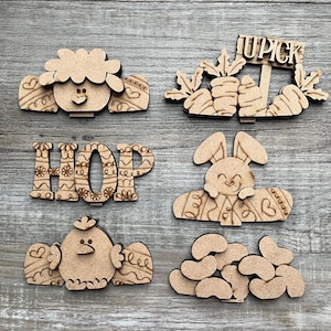 May include: A collection of laser-cut wooden Easter-themed decorations. Includes a sheep, a sign that says "UPICK" with carrots, the word "HOP", a bunny, a chick, and a pile of jelly beans. The decorations are light brown.