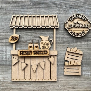 May include: A wooden lemonade stand with a sign that reads "FRESHLY SQUEEZED". The stand has a 25¢ sign, two drinks, and a pitcher. A sign that reads "FARMERS lemonade MARKET" is also included, along with a crate of lemons.
