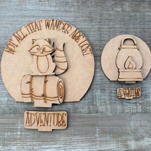 May include: Wooden camping-themed cutouts. One cutout features a raccoon on a rolled-up sleeping bag with the text "NOT ALL THAT WANDER ARE LOST". Another cutout shows a lantern with a flame, and a separate cutout that says "ADVENTURE".