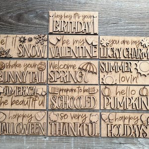 May include: Twelve wooden signs with various holiday and seasonal greetings. The signs are all in a rustic style with a light brown wood finish. The text on the signs includes "Happy Holidays", "Happy Halloween", "So Very Thankful", "Schooled", "Hello Pumpkin", "Summer Lovin", "Welcome Spring", "Shake Your Bunny Tail", "Let It Snow", "Be My Valentine", "You Are My Lucky Charm", and "Hey Hey It's Your Birthday".