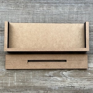 May include: Two unfinished wooden pieces for a DIY project. The top piece is a rectangular frame with a lip on the front edge. The bottom piece is a rectangular board with a slot cut out of the center.