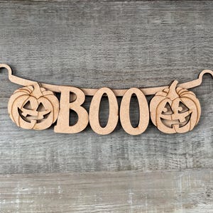 May include: A light brown wooden cutout banner featuring the word BOO in capital letters, flanked by two carved jack-o'-lantern pumpkins. The piece includes integrated hooks on each end for hanging, showcasing a flat, unfinished wood texture.