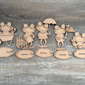 May include: A collection of wooden frog figurines, each with a unique design, including a frog with a wheelbarrow, a frog with a fence, a frog with an umbrella, a frog with a net, and a frog sitting down. Each frog is placed on a wooden lily pad.