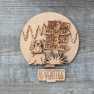 May include: Wooden sign with a circular top featuring a raccoon, trees, and directional arrows pointing to hiking trails, bathrooms, picnic area, and fishing pond. The word "ADVENTURE" is displayed below. The sign is made of light brown wood.