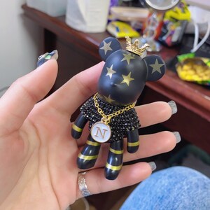 May include: Black and gold bear figurine with a gold crown and a gold chain with a white circle pendant with the letter "N" on it. The bear has gold stars on its body.