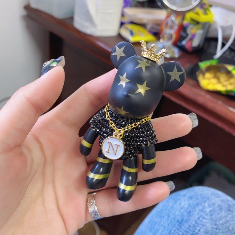 May include: Black and gold bear figurine with a gold crown and a gold chain with a white circle pendant with the letter "N" on it. The bear has gold stars on its body.