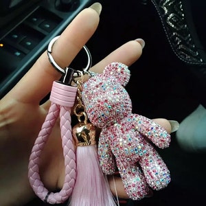 May include: A pink and silver rhinestone teddy bear keychain with a pink braided wrist strap and tassel. The bear is covered in sparkling rhinestones and has a gold-colored bell charm.