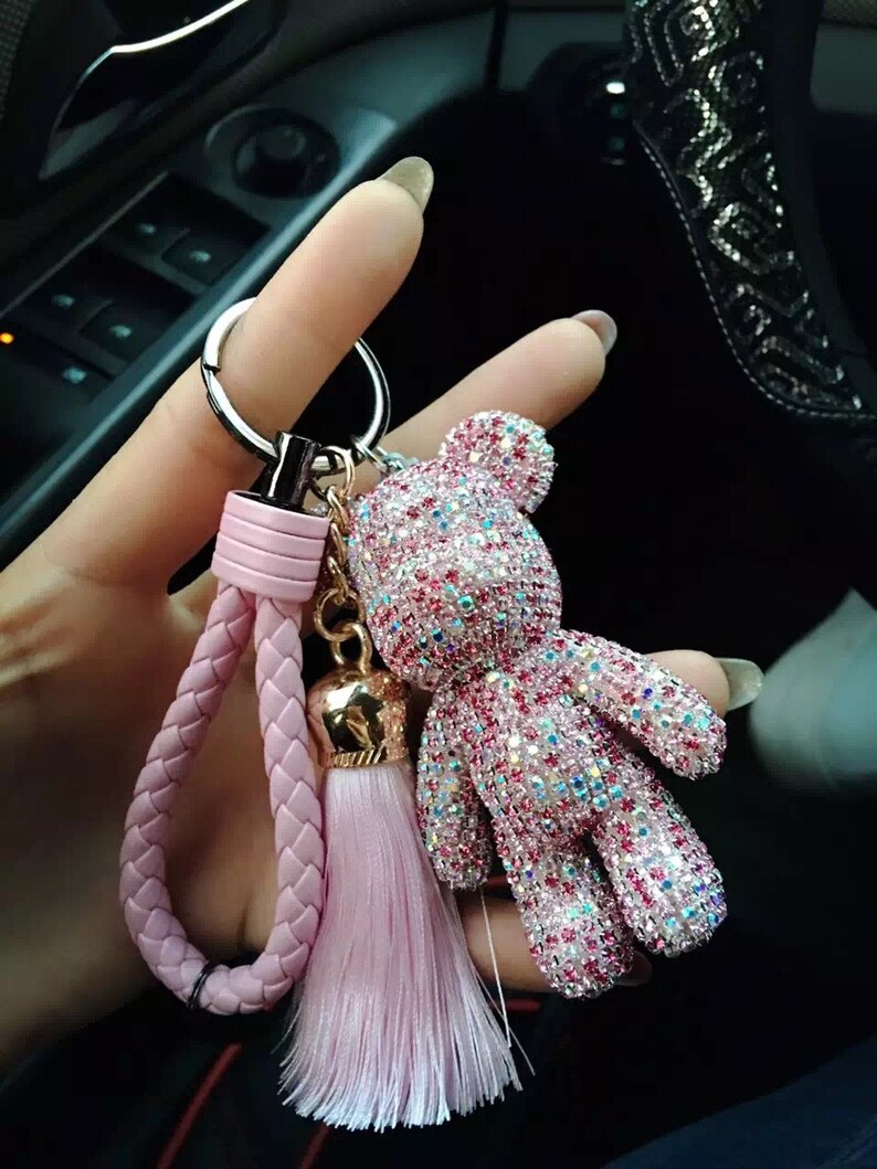 May include: A pink and silver rhinestone teddy bear keychain with a pink braided wrist strap and tassel. The bear is covered in sparkling rhinestones and has a gold-colored bell charm.