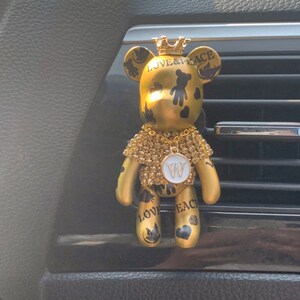 May include: Gold-colored teddy bear car air freshener with a crown on its head. The bear is decorated with black and white details and the words "LOVE & PEACE". The bear is wearing a gold chain with a white letter "W" pendant.