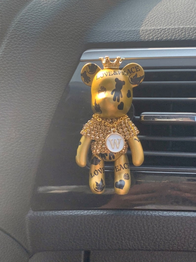 May include: Gold-colored teddy bear car air freshener with a crown on its head. The bear is decorated with black and white details and the words "LOVE & PEACE". The bear is wearing a gold chain with a white letter "W" pendant.