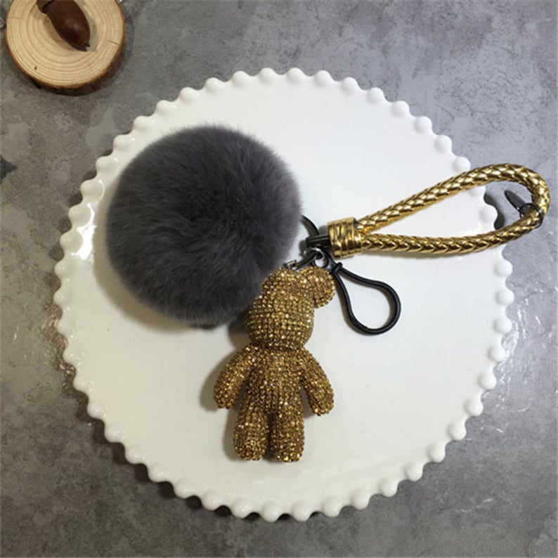 May include: A gold rhinestone teddy bear keychain with a gray fluffy pom-pom attached to a gold chain.