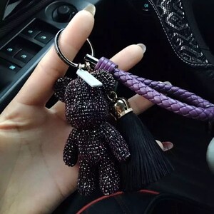 May include: A purple jeweled teddy bear keychain with a black tassel and a braided purple strap. The keychain is attached to a silver ring. The bear is covered in small, sparkling purple gems.