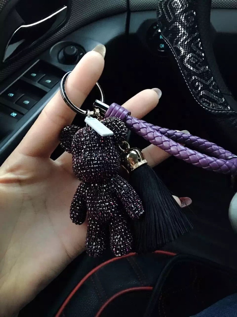 May include: A purple jeweled teddy bear keychain with a black tassel and a braided purple strap. The keychain is attached to a silver ring. The bear is covered in small, sparkling purple gems.