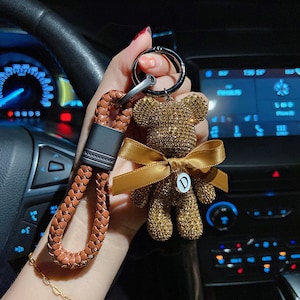 Könnte beinhalten: Ein goldfarbener Strass-Teddybär-Schlüsselanhänger mit braunem Band und einem Buchstaben "D"-Anhänger. Der Schlüsselanhänger hat einen braunen geflochtenen Lederriemen und einen silbernen Metallring.