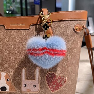 May include: A light blue and red heart-shaped fur keychain with a gold chain and black flower charm attached to a brown leather tote bag with a white and brown patterned design.