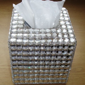 Puede incluir: Una tapa de caja de pañuelos cuadrada con un diseño de cristal plateado y transparente. La tapa está hecha de pequeños cristales cuadrados que están dispuestos en un patrón de cuadrícula.