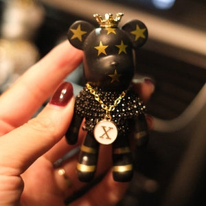 May include: Black bear figurine with gold stars and a gold crown. The bear is wearing a black beaded necklace with a gold chain and a gold pendant with the letter 'X' on it.