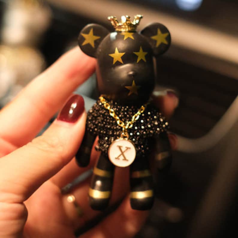 May include: Black bear figurine with gold stars and a gold crown. The bear is wearing a black beaded necklace with a gold chain and a gold pendant with the letter 'X' on it.